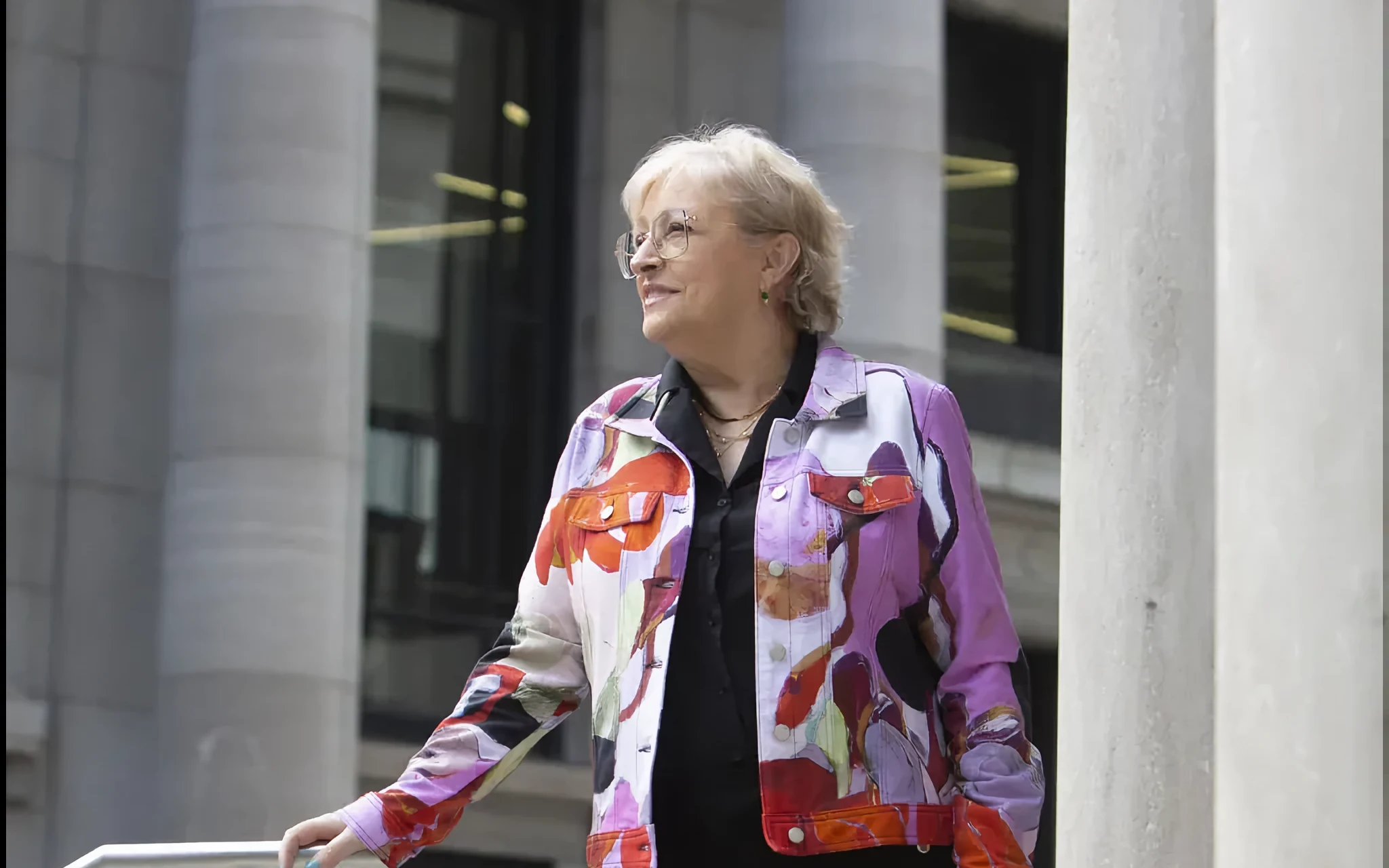Christiane Constantineau, souriante, en veste à motifs colorés, appuyée à une rampe devant des colonnes de pierre