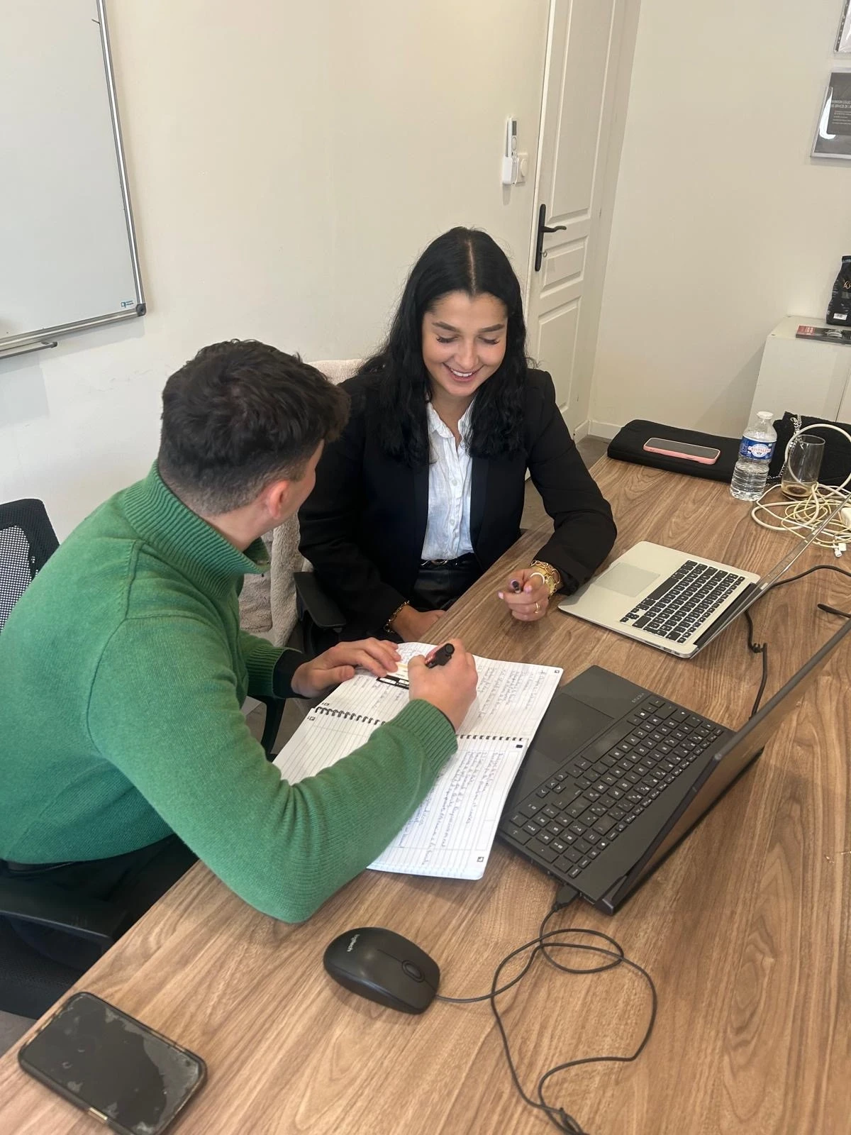 Un homme et une femme souriant. Ils sont assis autour d'une table avec un ordinateur portable et écrivent sur un cahier.