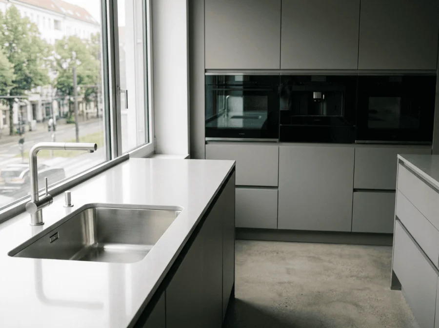 Eine moderne, minimalistische Küche in Grautönen mit einer weißen Arbeitsinsel, Spülbecken aus Edelstahl, großen Fenstern zur Straße und einer Küchenzeile mit eingebauten Elektrogeräten im Hintergrund.
