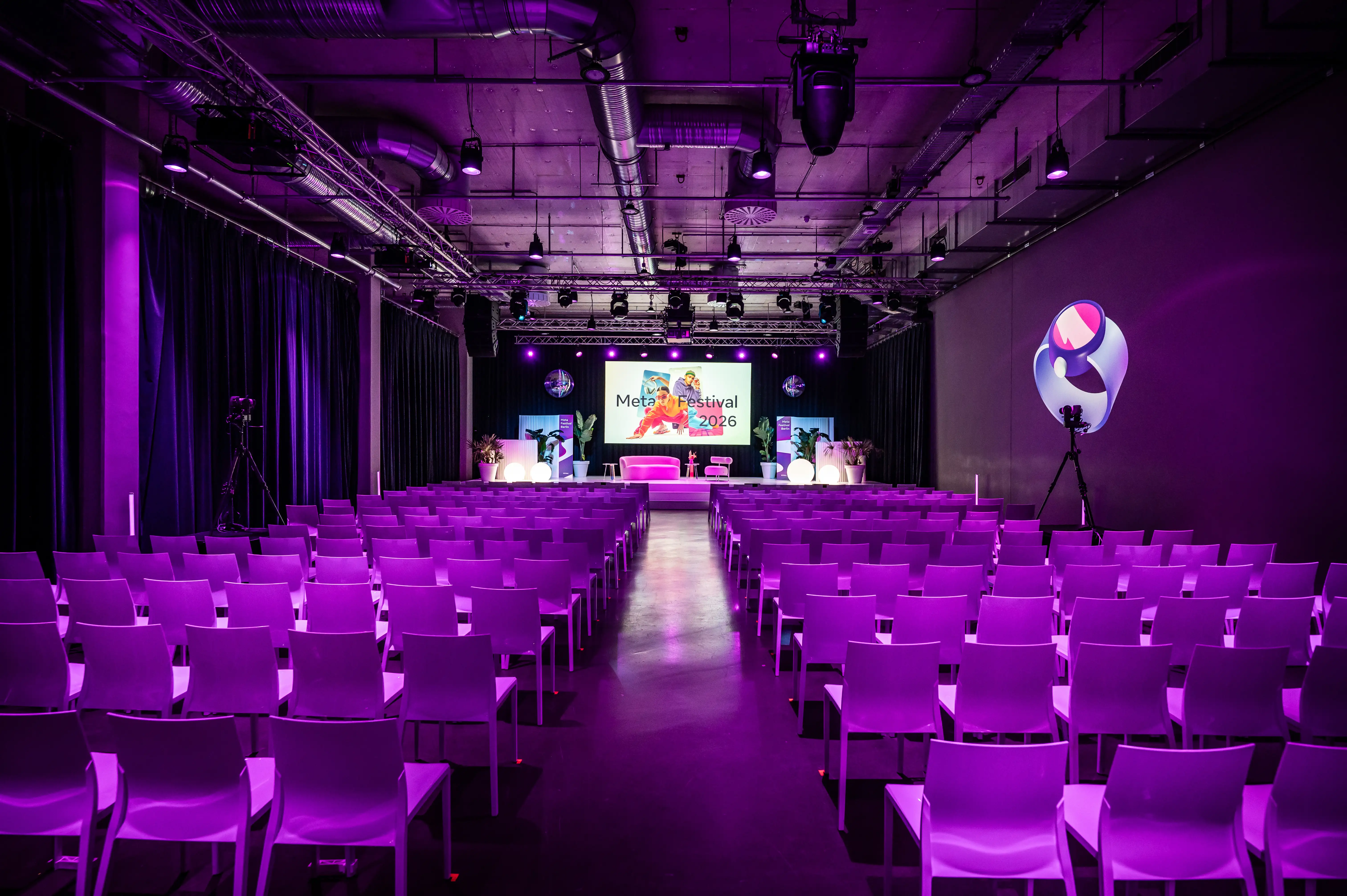 Purple-lit hall with rows of white chairs facing the Meta Festival stage and disco balls.