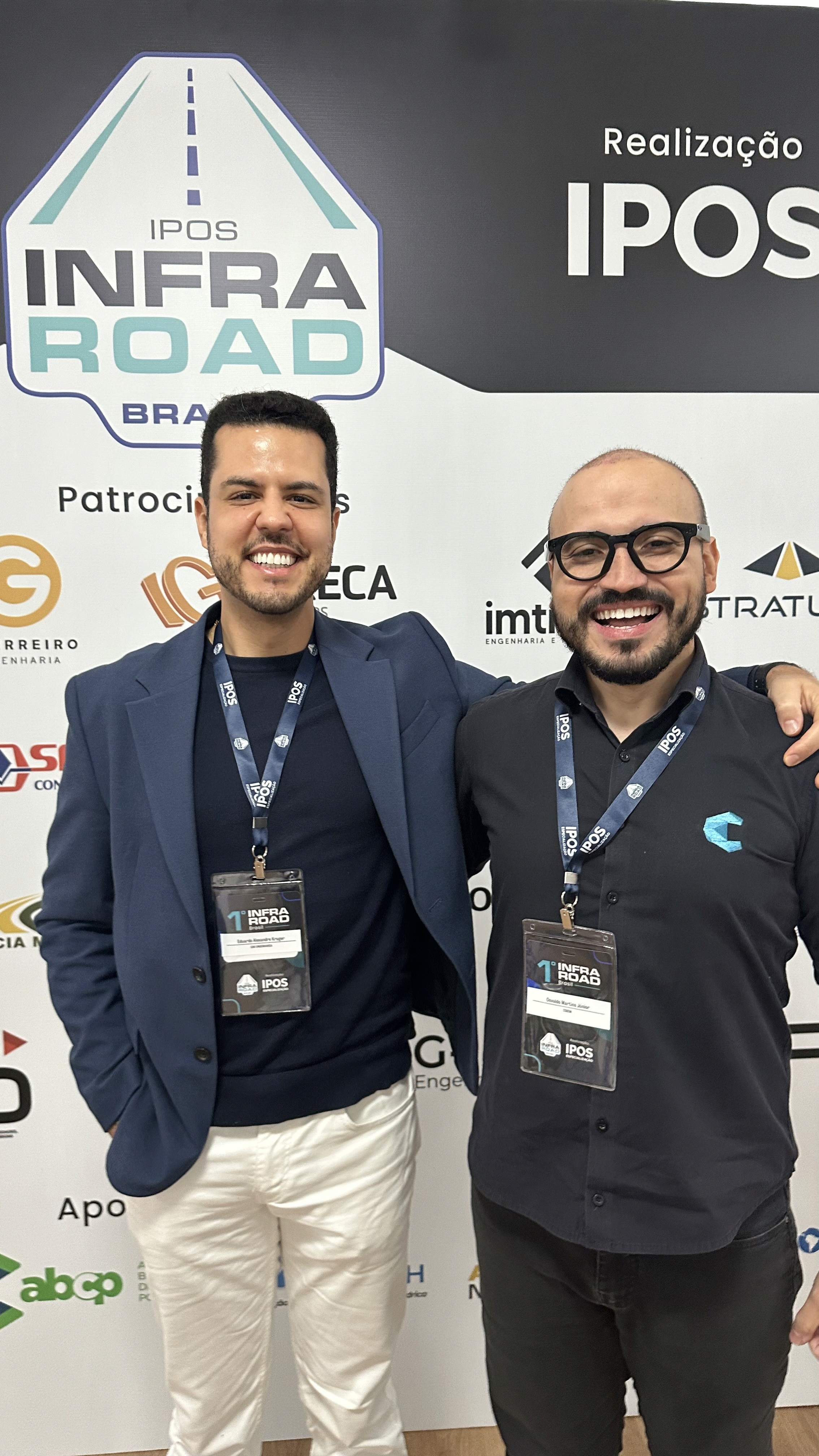 Eduardo Alexandre Kruger e colega sorrindo com credenciais do evento IPOS Infra Road Brasil, focado em engenharia e infraestrutura rodoviária.