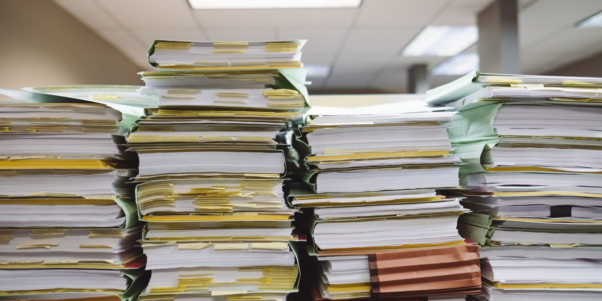 A large stack of organized paperwork and files is neatly arranged on a desk in a well-lit office environment, highlighting the themes of organization, administration, and office work.