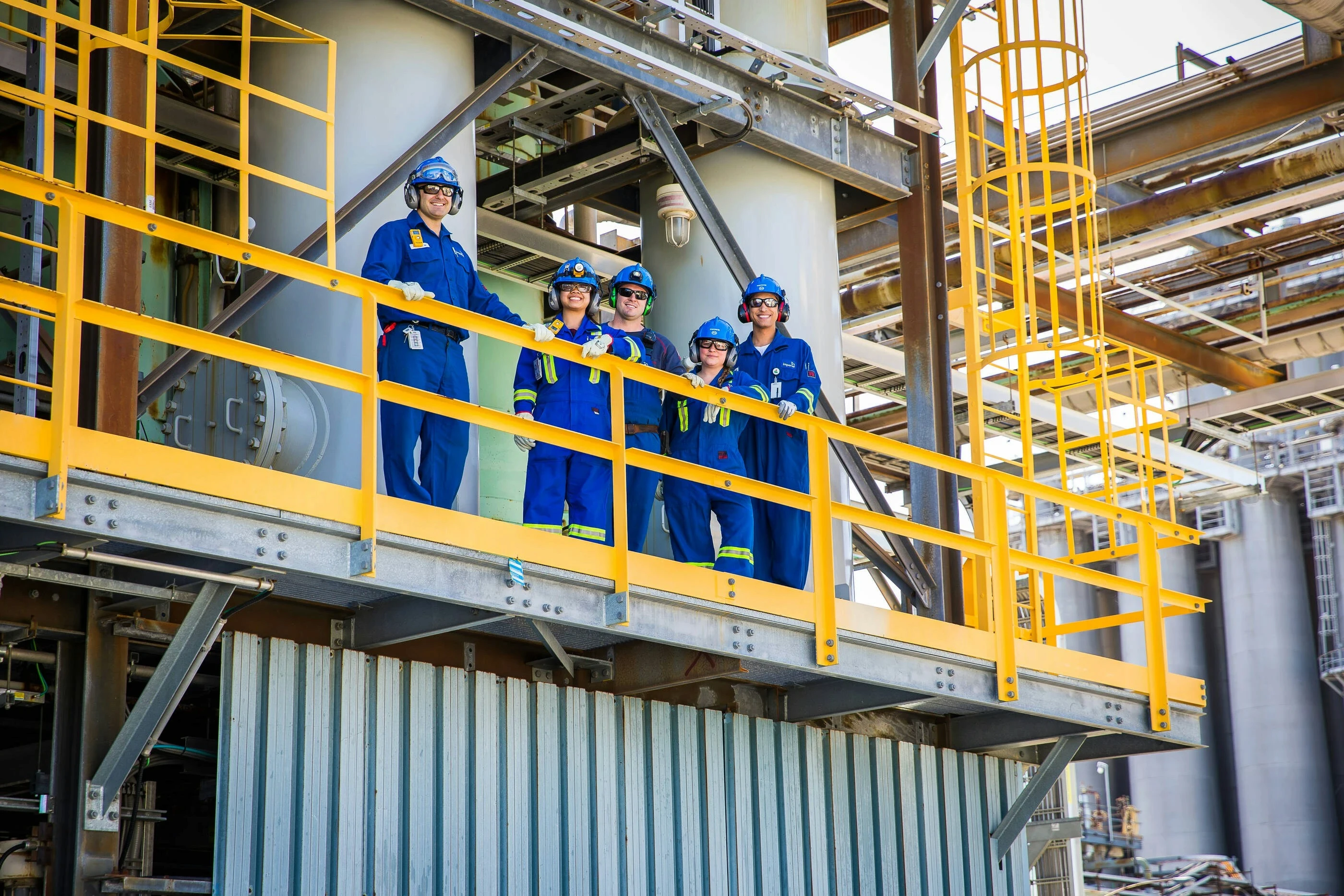Five industrial workers in blue coveralls and PPE standing on a yellow-railed platform.