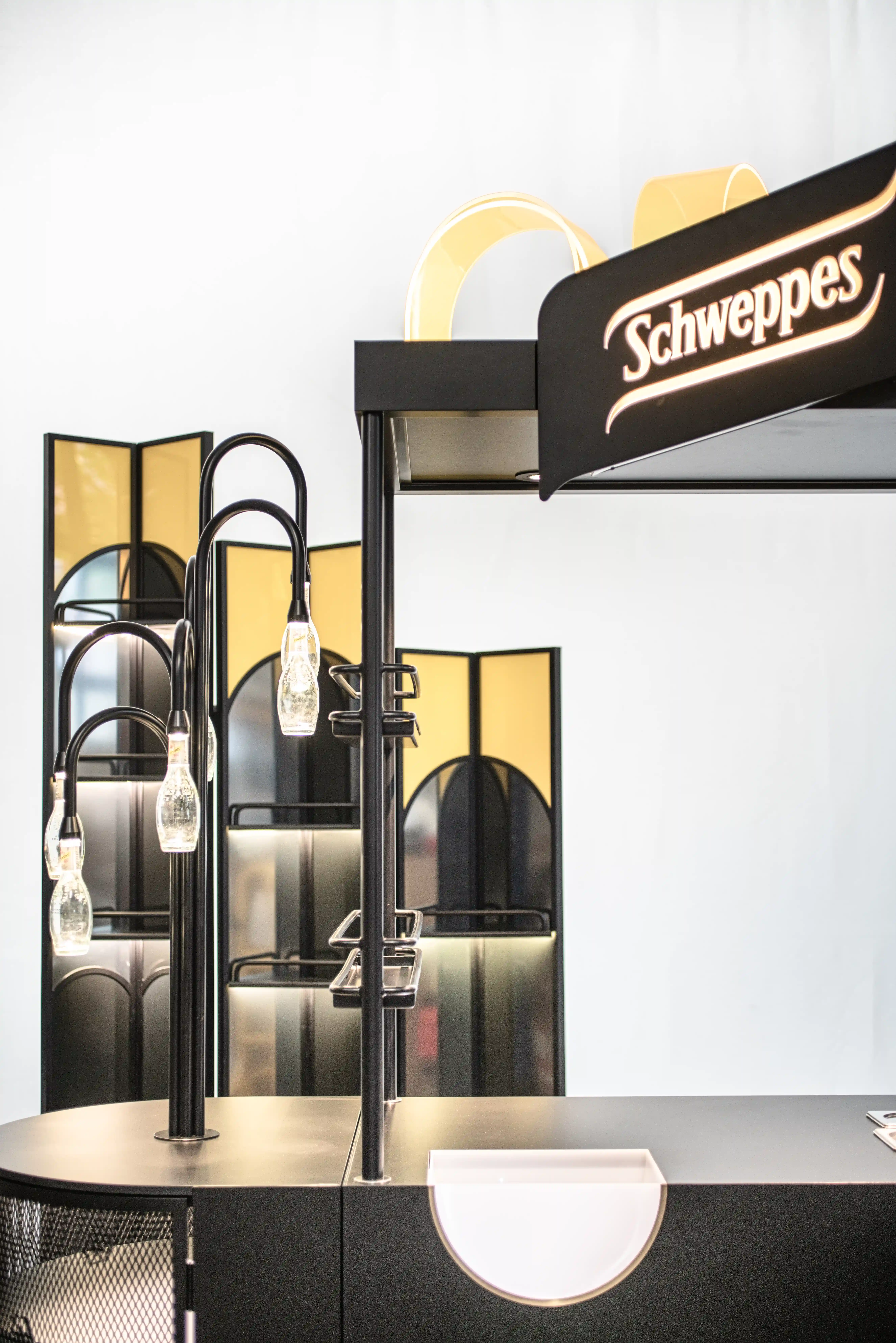 Detail of the SCHWEPPES bartool with an illuminated black logo sign, golden arch elements and curved black lamp arms holding bottle-shaped lights.