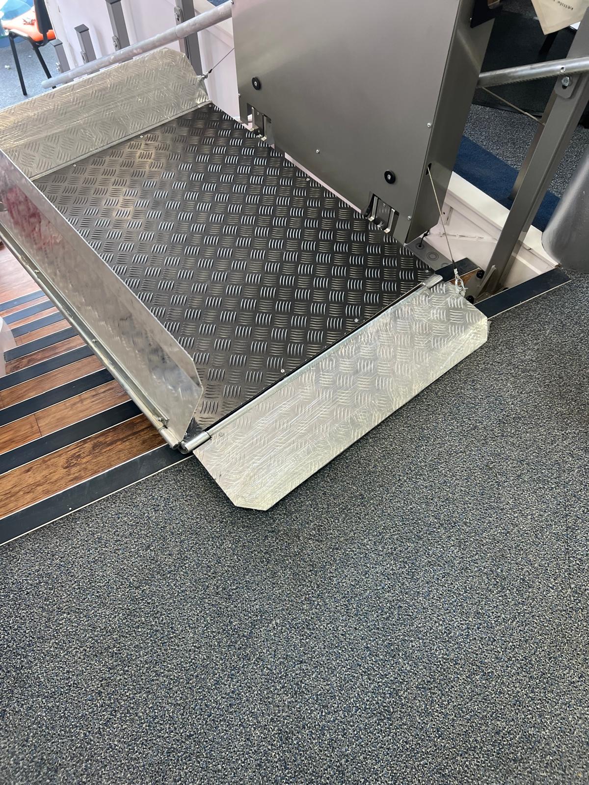 Indoor inclined platform stairlift at lower landing — anti-slip chequerplate platform with folding entry ramp deployed onto carpeted floor, staircase rail visible to the left, commercial interior installation