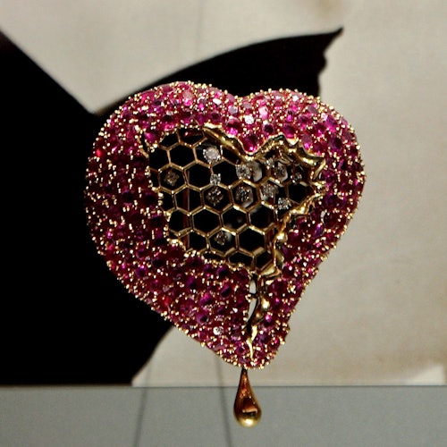 יצירת אמנות בצורת לב מעוטרת באבני חן ורודות, עם עיצוב רשת משושים ואלמנט זהב בצורת דמעה בתחתית.