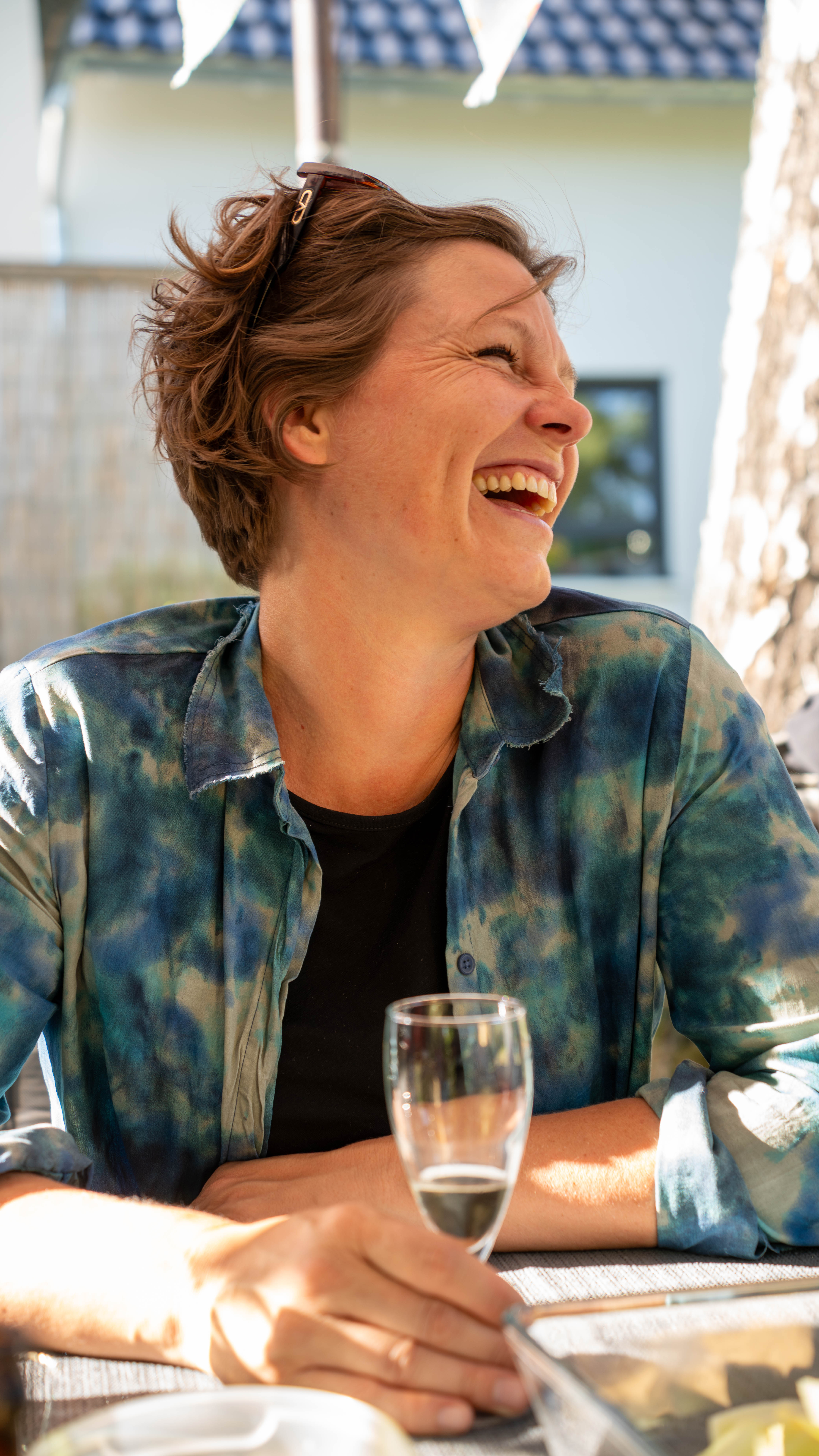 Frau mit kurz geschnittenem Haar und blauer Batikbluse sitzt an einem Tisch im Freien und hält ein Glas mit Getränk