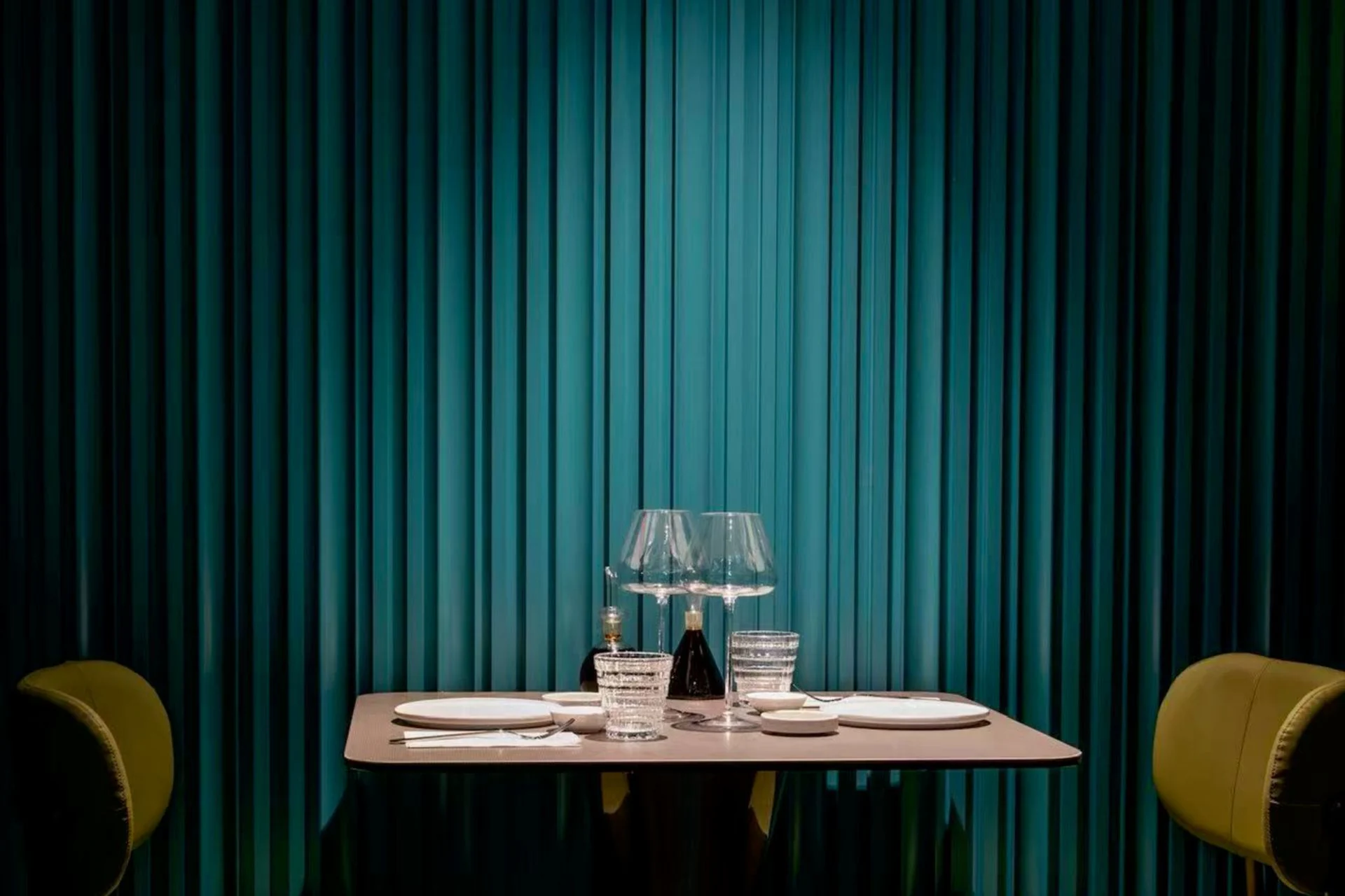 Table dressée sous un projecteur, entre deux pans de rideau de velours bleu canard et deux fauteuils olive