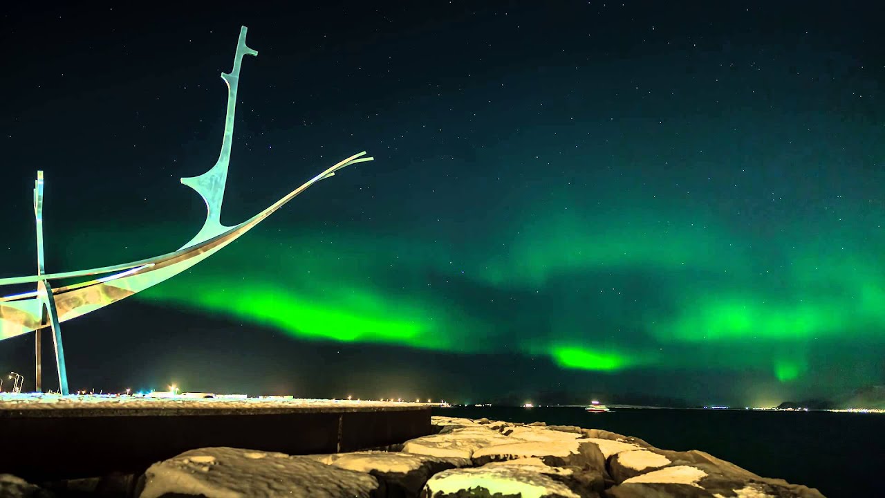 Faint green aurora arcing over open ground with the low Reykjavik skyline on the horizon in winter