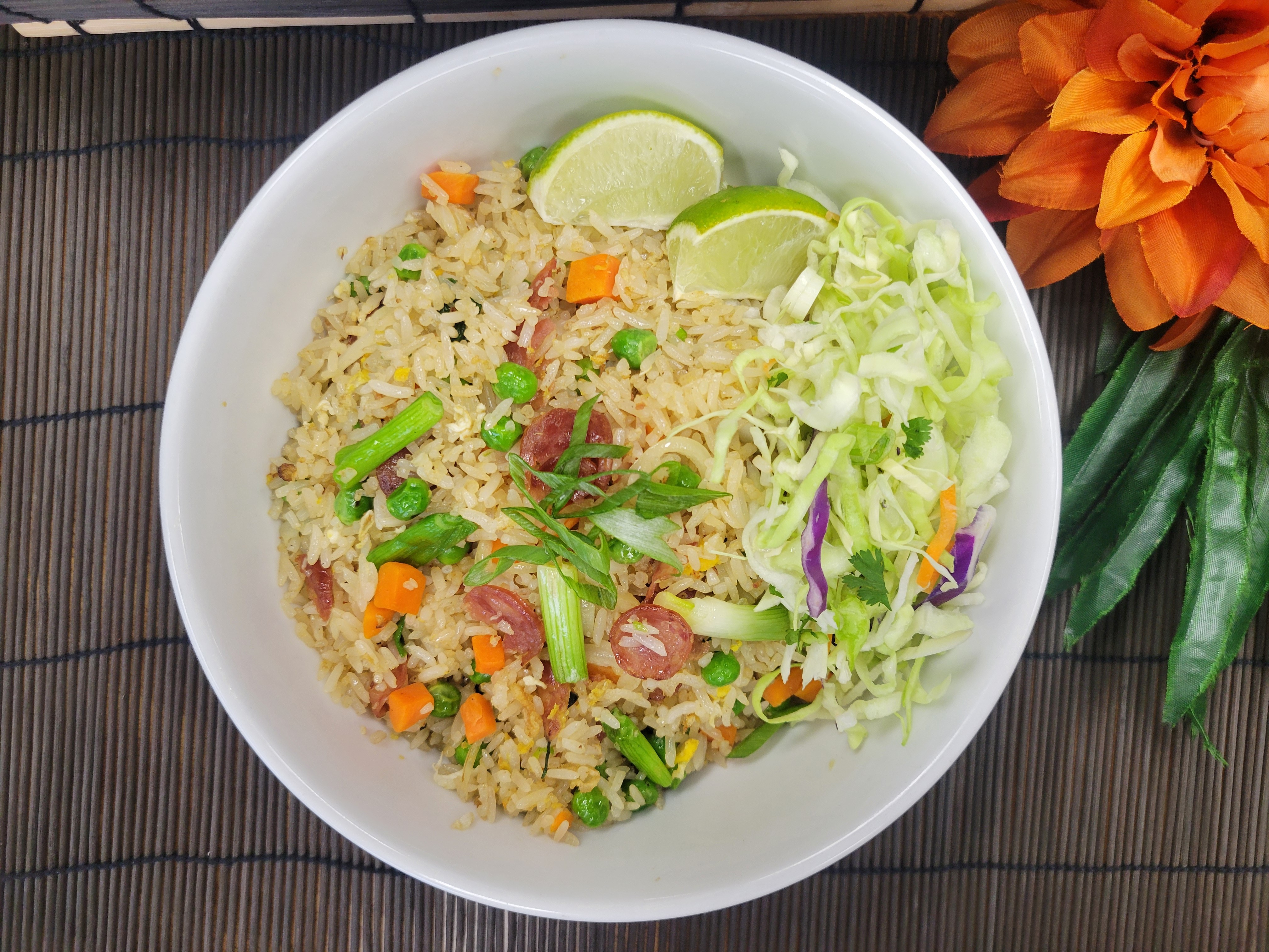 A white bowl filled with garlic fried rice containing peas, carrots, and slices of Chinese sausage, garnished with green onions and served with lime wedges and Asian slaw.