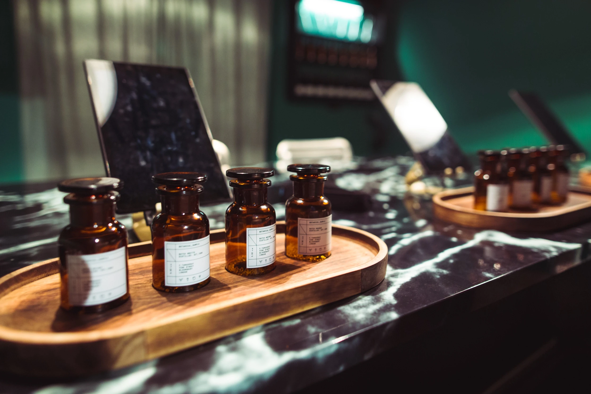 Apothecary-style tasting bottles with paper labels on wooden trays along a black marble counter at DIAGEO BCB18.