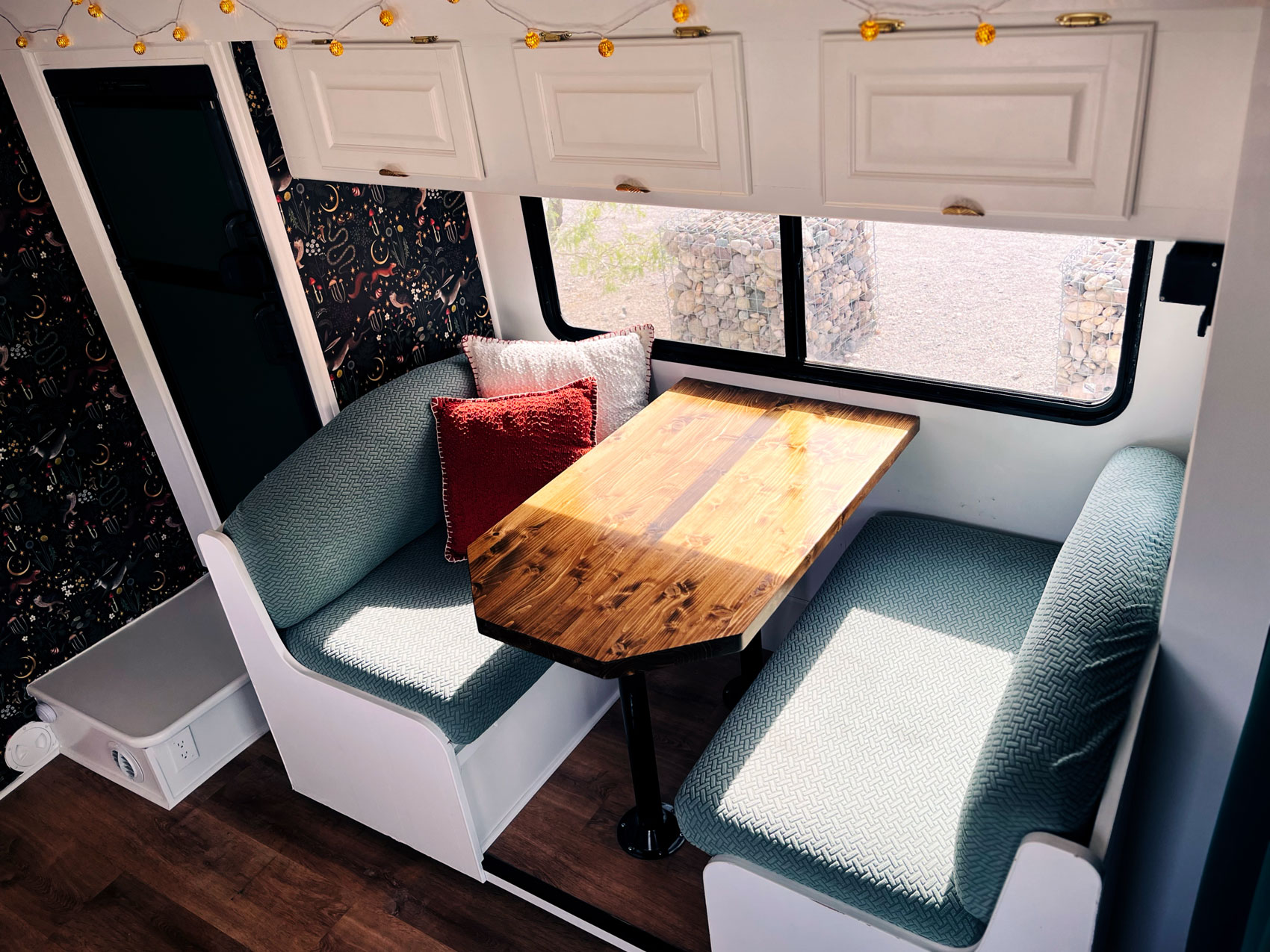 Cozy RV dining nook with a wooden table and teal cushioned bench seats. White cabinets above, with string lights adding a warm ambiance.