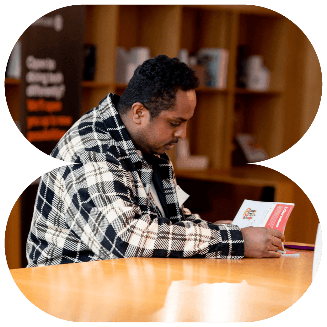 Person sitting at a wooden table reading a booklet, wearing a patterned check coat in a room with bookshelves.