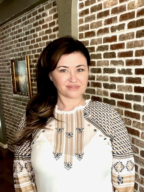 Woman with long brown hair stands smiling in front of a brick wall. She wears a white blouse with patterned sleeves and detailing. A framed painting hangs on the wall behind her.