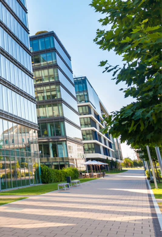 Modern glass buildings line a walkway, surrounded by greenery and clear blue skies on a sunny day.