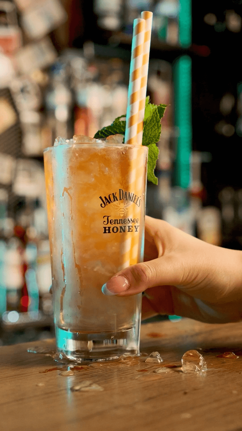 Fotografia kulinarna przedstawiająca zbliżenie na rękę trzymającą żółty drink z dwiema słomkami w barze