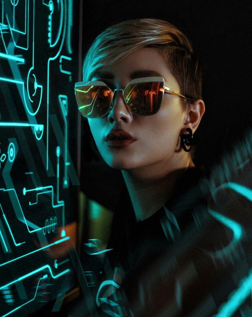 A stylish individual with short hair and reflective sunglasses stands in a futuristic setting illuminated by neon lights and digital interface graphics. The person's edgy look and the high-tech background evoke a cyberpunk aesthetic, emphasizing themes of technology, modernity, and futuristic fashion.