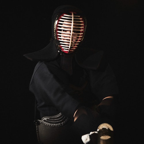 Een persoon gekleed in kendo-uitrusting, inclusief een beschermend gezichtsmasker met een gerasterd ontwerp, die een kendo-stok vasthoudt in een zwak verlichte omgeving.