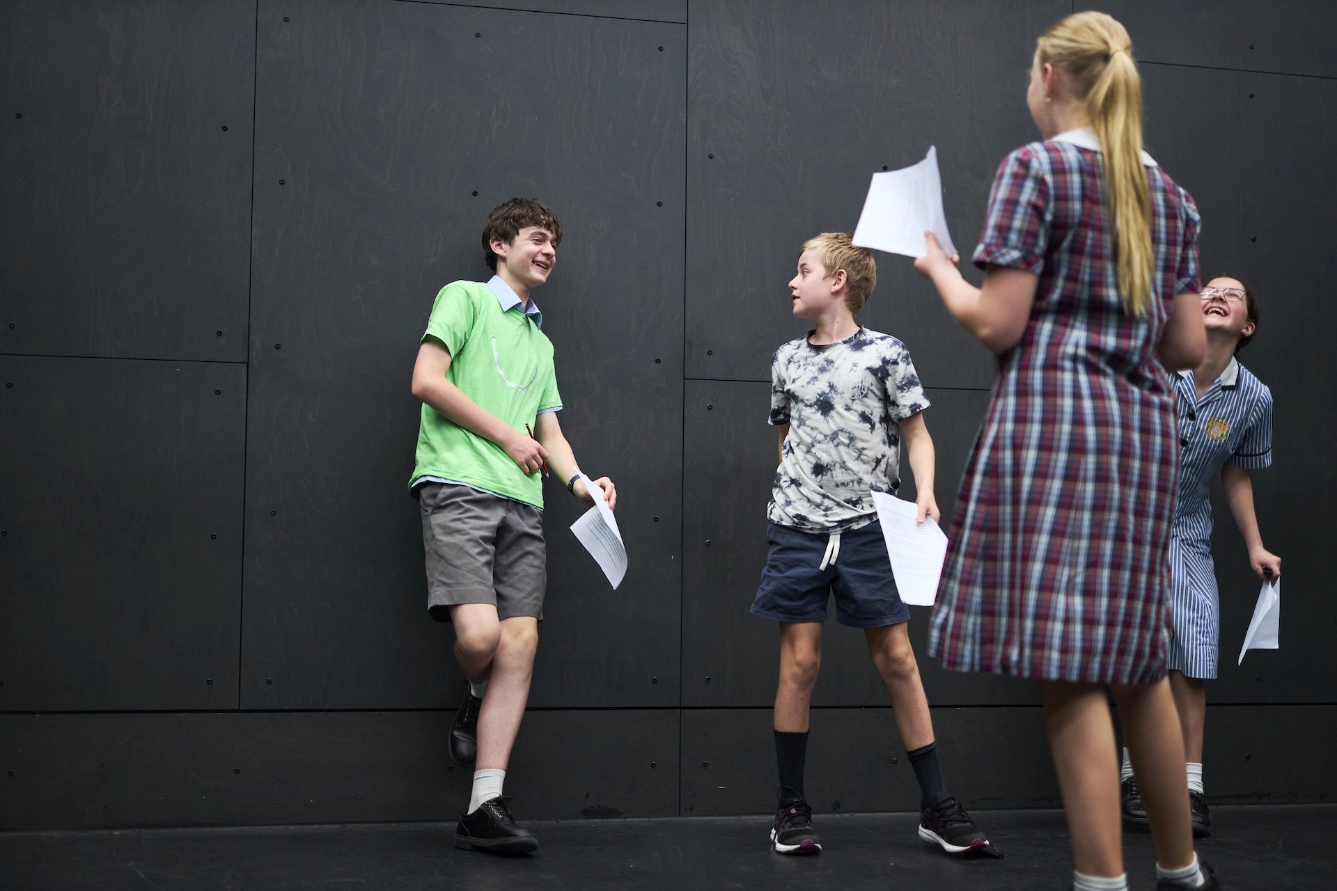 Four young performers rehearse a scene in a studio, laughing and holding scripts while standing against a dark wall.
