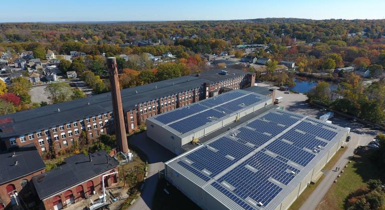 Aerial view of a building with solar panels on the roof, surrounded by trees and open landscape.