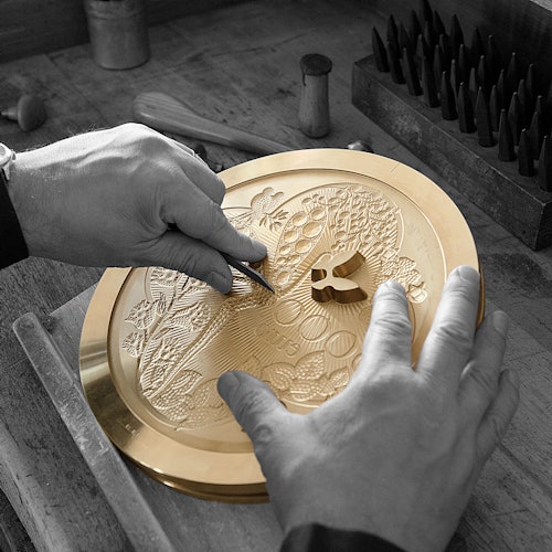 在一个木制工作台上，一双手正用雕刻工具在圆形黄铜板上进行细致的雕刻，周围摆放着几种雕刻工具。
