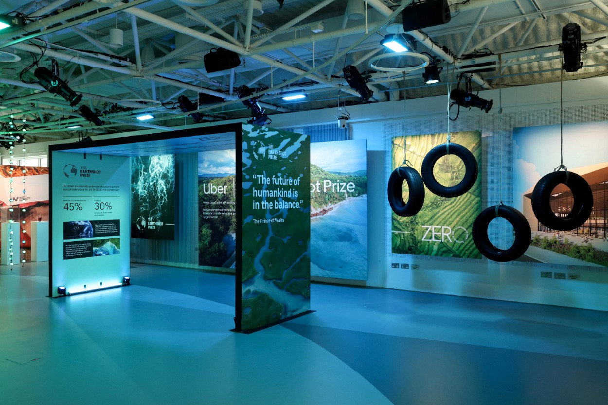 An immersive exhibition space with cool blue-green lighting and an exposed industrial ceiling rig. Large freestanding display panels feature landscape photography, data, and the quote "The future of humankind is in the balance." Hanging tyre installations add a sculptural element to the space.