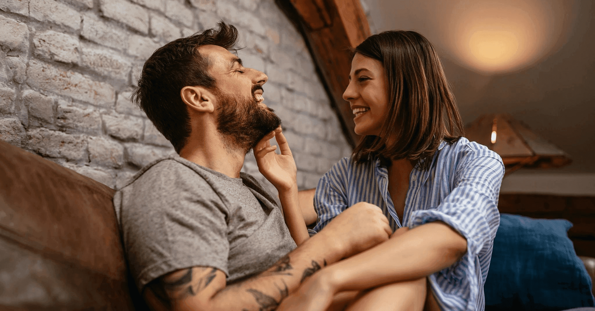 Ett lyckligt par sitter nära varandra i ett mysigt vardagsrum medan de delar ett ömt ögonblick och ler in i varandras ögon, vilket illustrerar närhet och kärlek i en modern relation.