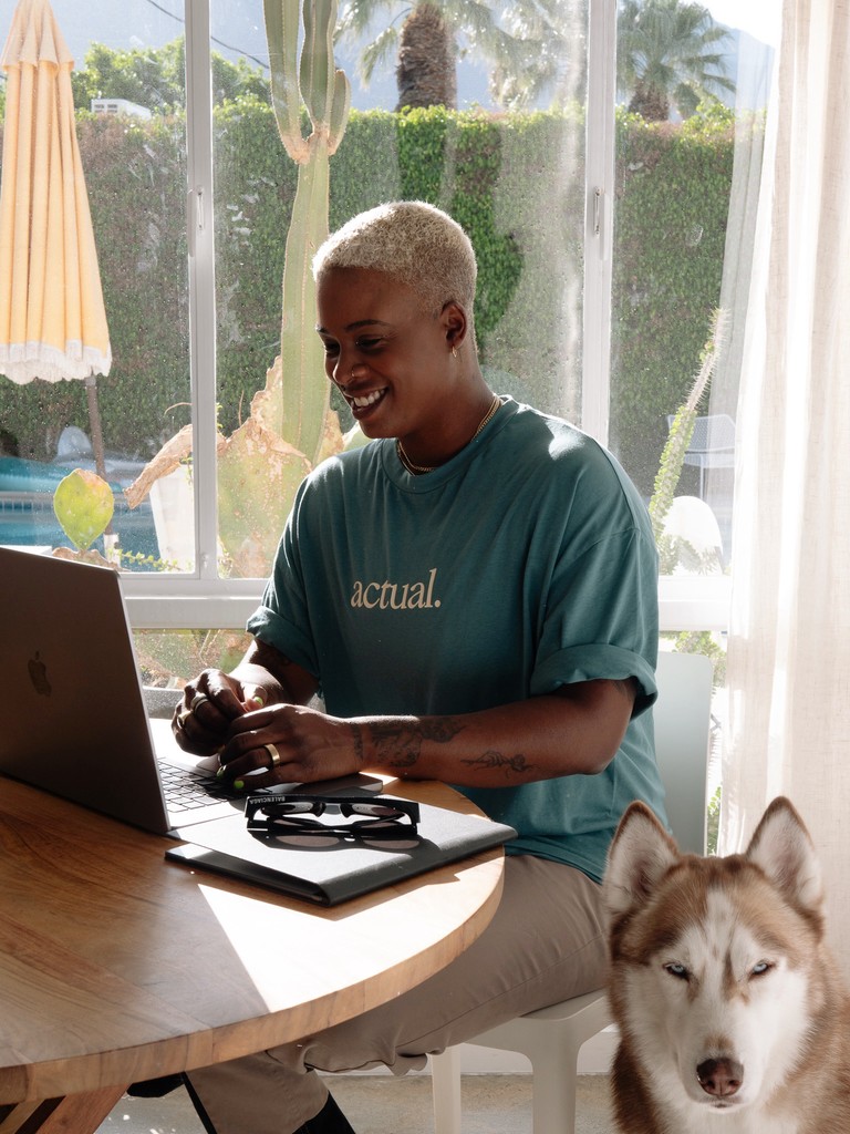 Ashleigh Warren sitting at a table, working on a laptop, with a dog lying nearby and sunlight streaming in through the window.