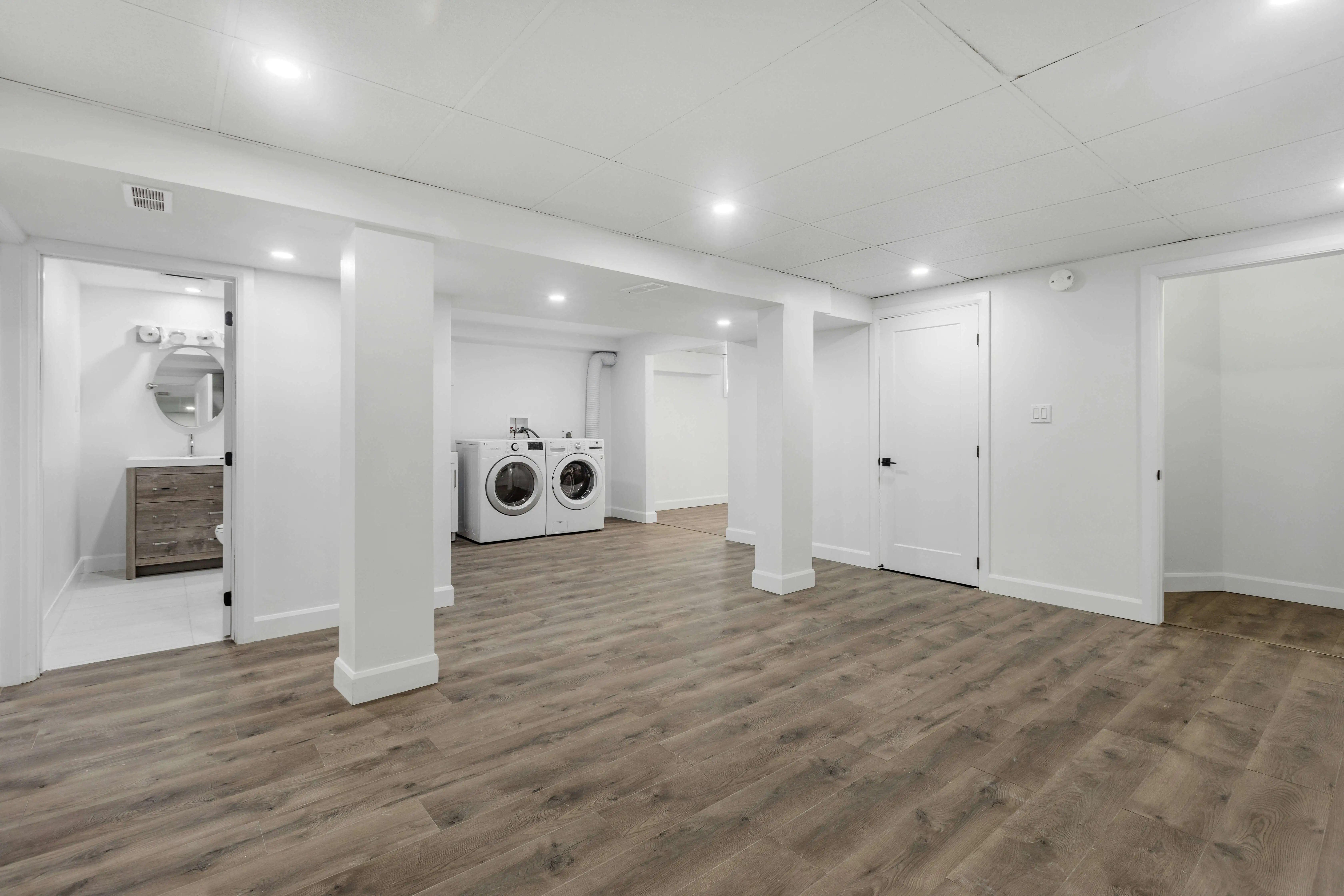 Spacious basement with wood flooring, bright recessed lighting, a laundry area with washer and dryer, white walls, and an adjacent bathroom. Clean and modern.