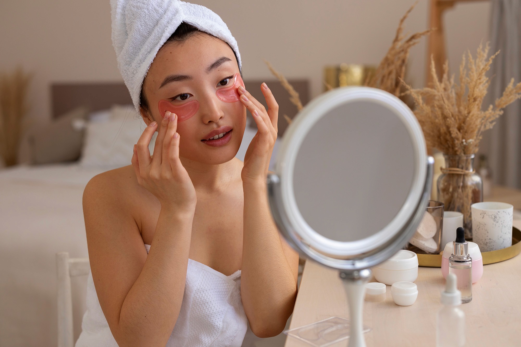 Woman in a towel applying pink under-eye gel patches while looking into a mirror on a vanity table