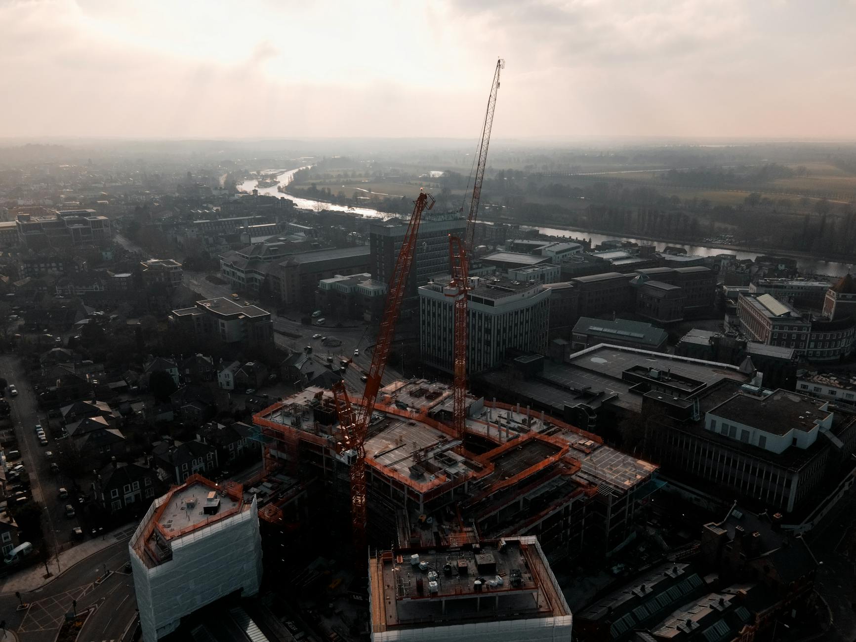 Aerial view of a city construction site with a tall orange tower crane, surrounding rooftops and a river at dawn.