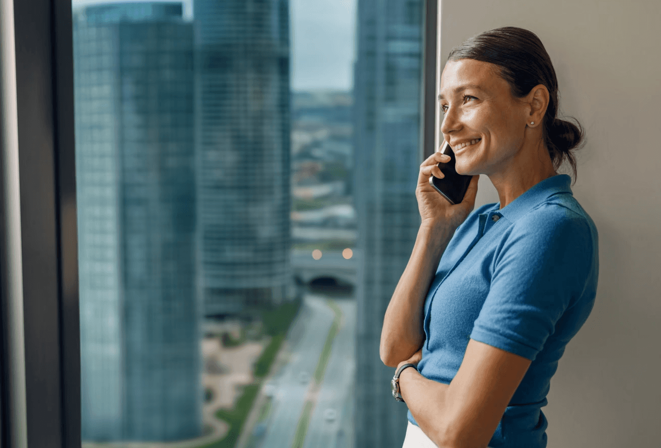 A business professional in a blue shirt smiles while talking on a smartphone, standing by a window with a view of modern city skyscrapers.