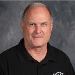 A professional headshot of an American man in his fifties wearing a black polo shirt with a school logo on it, against a neutral background.