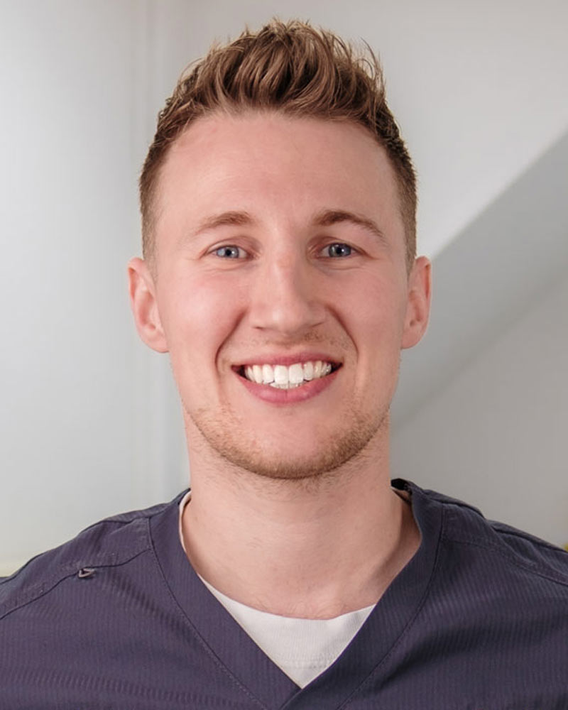 A portrait of Harvey, an Associate Dentist at Cricklade Dental Practice, smiling and wearing a dark grey scrub top with yellow embroidered text that reads "Harvey" on the left and "Cricklade Dental Practice" on the right. He is standing in a dental surgery with a clock and equipment visible in the background.