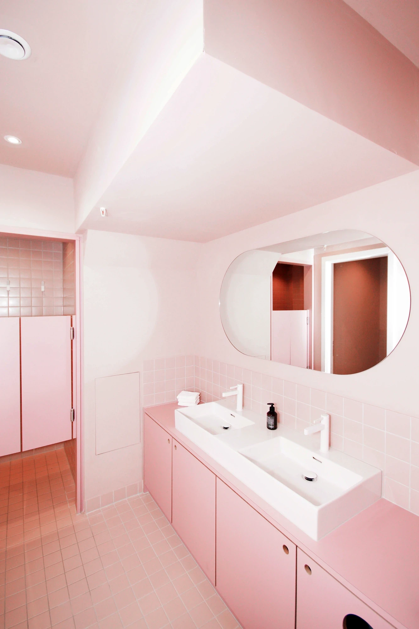 Pastel pink bathroom interior with double white sinks on a pink countertop, oval mirror, pink tiled walls and floor.