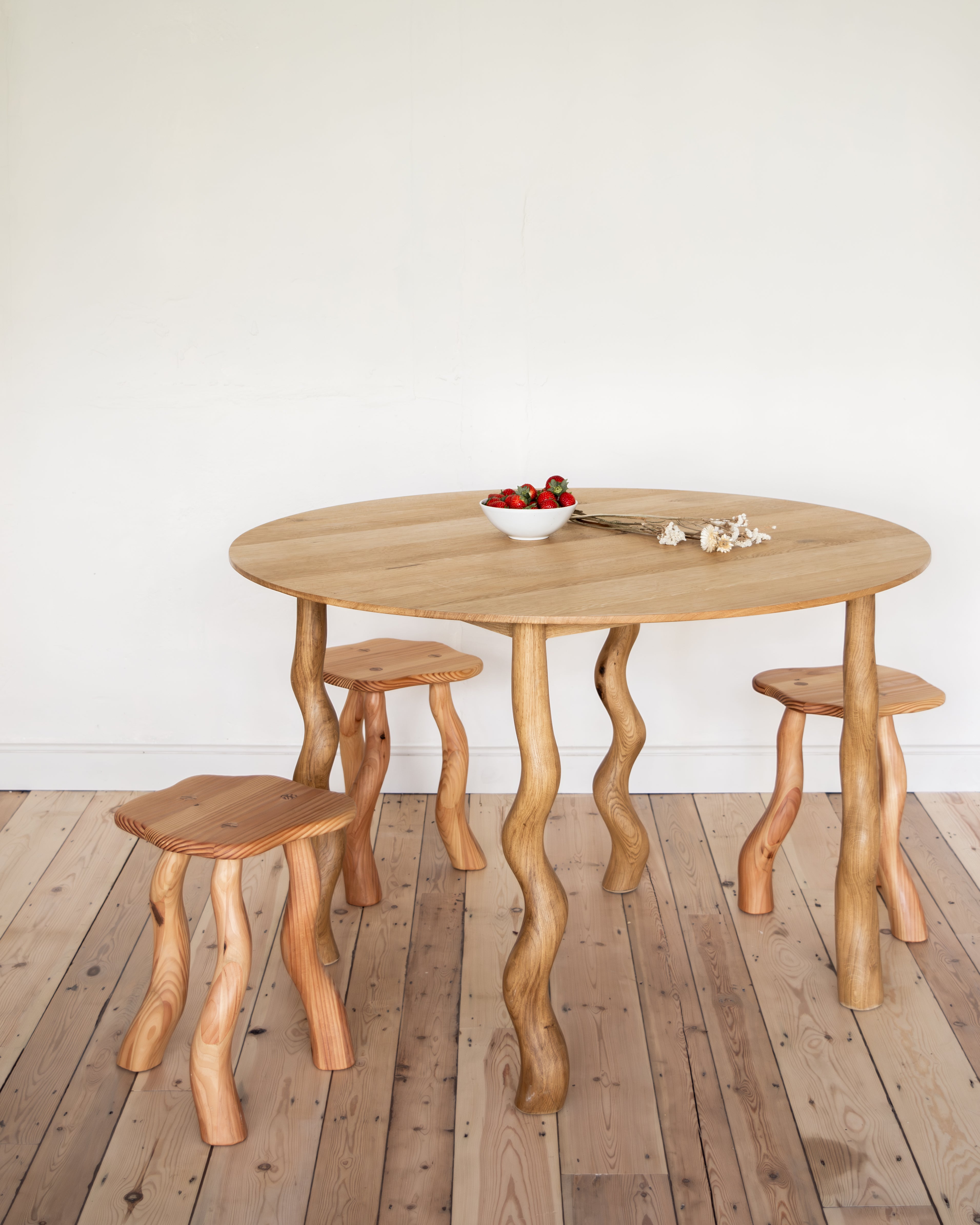 Wooden dining table with three stools on a wooden floor against a white wall.