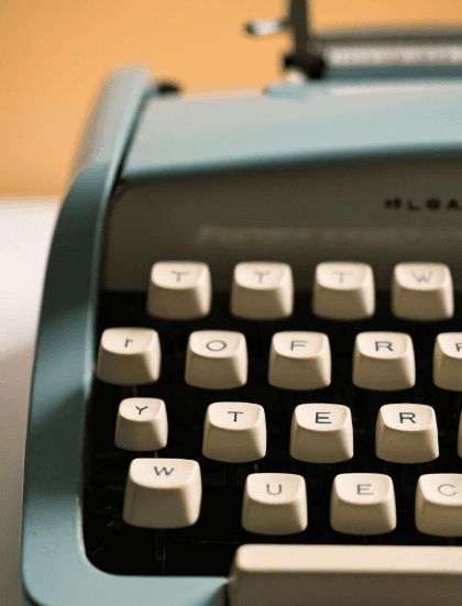 Close-up of a vintage blue typewriter with white keys, angled slightly to the left, against a warm beige background. The image evokes nostalgia.