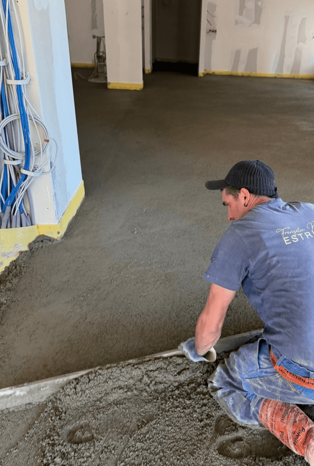 Ein Bauarbeiter, der Arbeitskleidung und eine Kopfbedeckung trägt, ebnet mit einer langen Abziehlatte einen frisch betonierten Boden.
