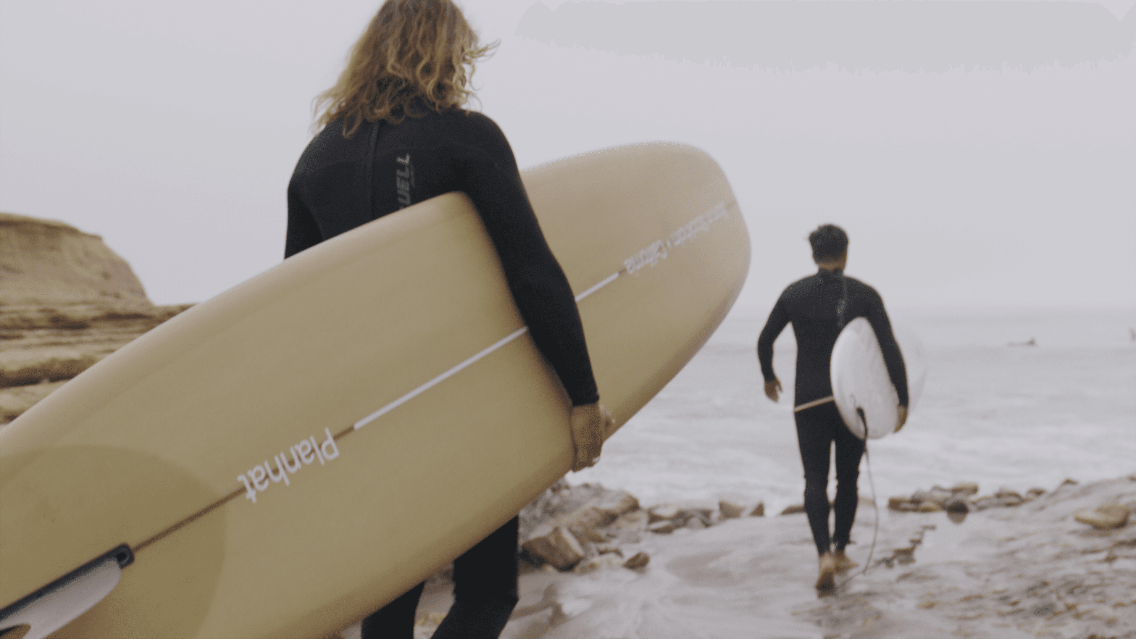 Two surfers in black wetsuits walk toward the ocean on an overcast day, carrying surfboards — one tan with Planhat branding, one white.