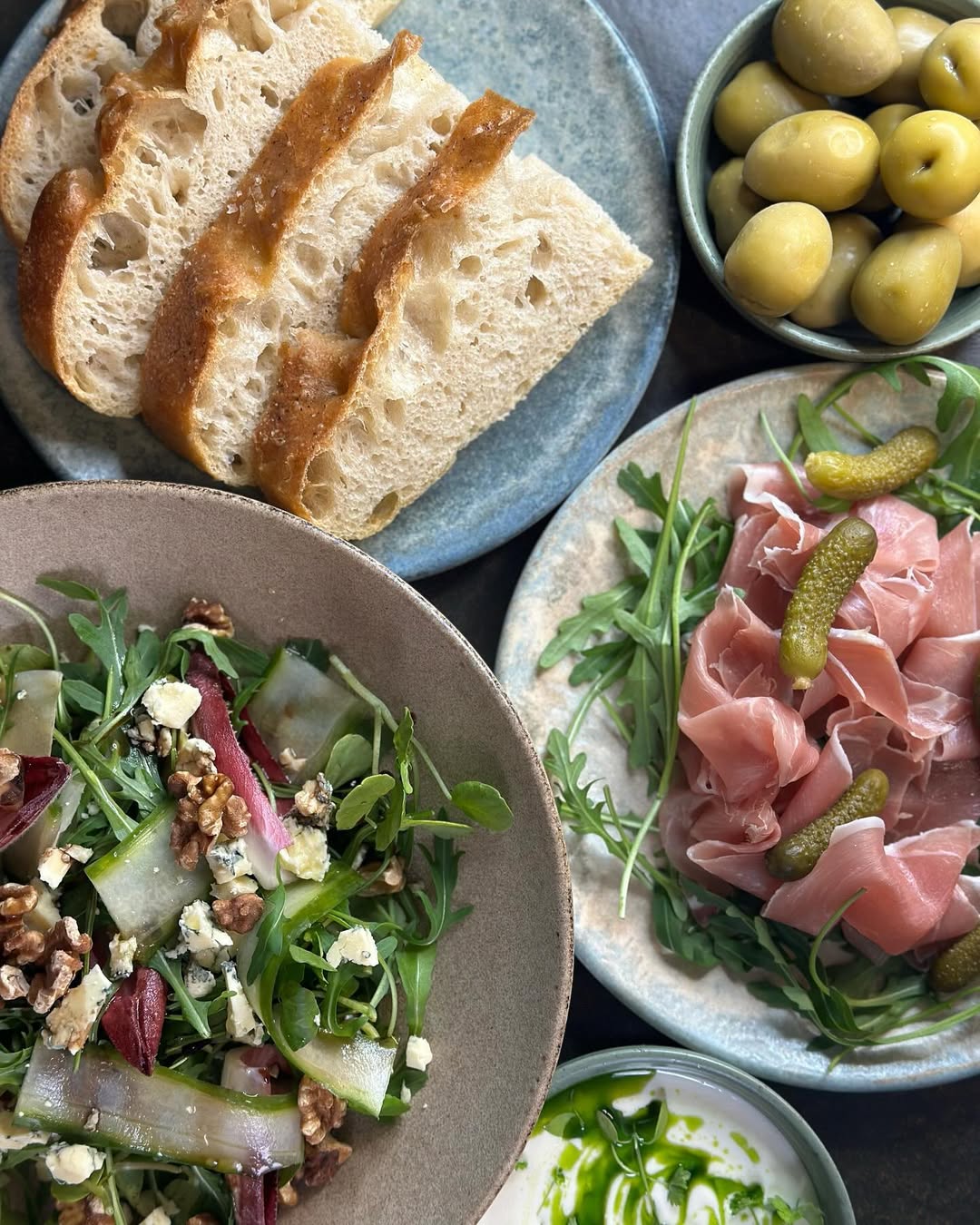 Variety of dishes: bread slices, potatoes, salad, prosciutto with pickles, and dip, for a delicious meal.