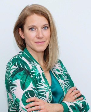 A woman with blonde hair wearing a green and white patterned blazer smiles confidently with arms crossed.