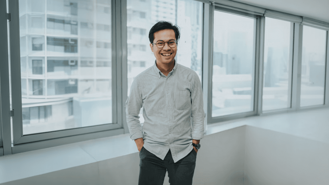 A smiling man stands casually in an office environment, wearing glasses, a light striped shirt, and dark pants. His hands rest in his pockets, conveying a relaxed demeanor. Behind him, large windows reveal an urban cityscape, with buildings visible in the background. The lighting is soft and natural, casting gentle highlights and creating a calm atmosphere. The composition is centered, capturing the subject from a straight-on perspective. The mood is professional yet approachable.