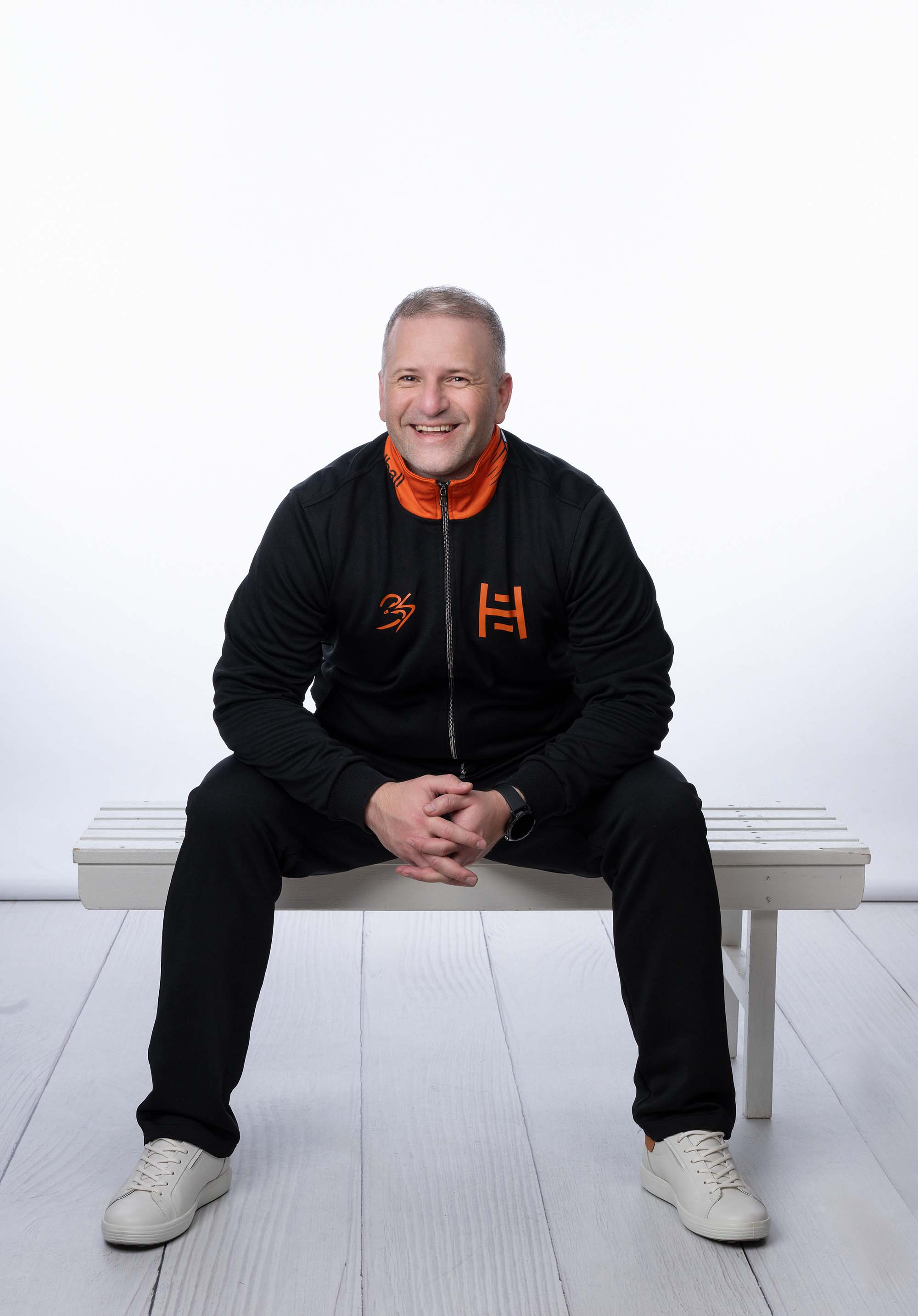 A coach sitting on a white bench wearing a black and orange tracksuit.