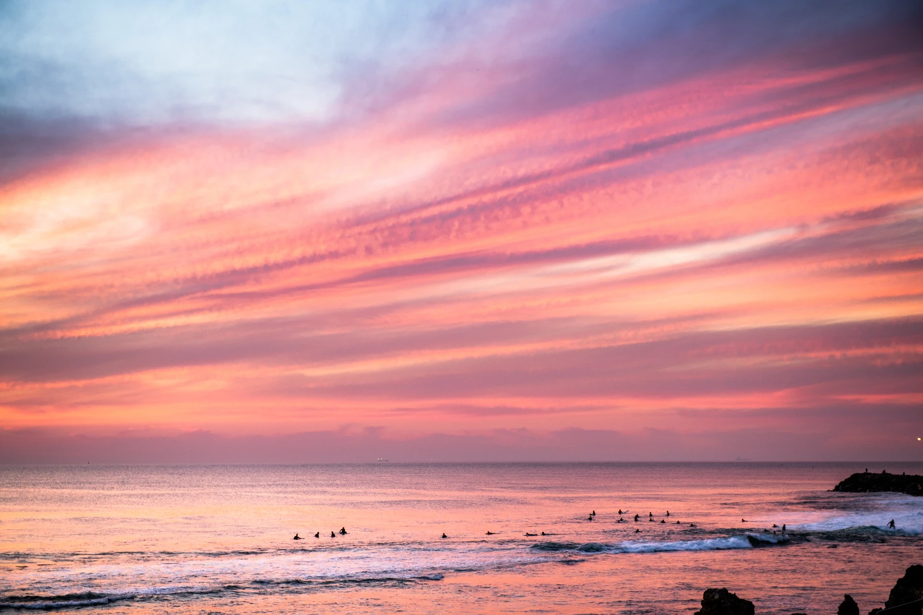 Surfers waiting in the line-up beneath a streaked pink and orange sunset sky
