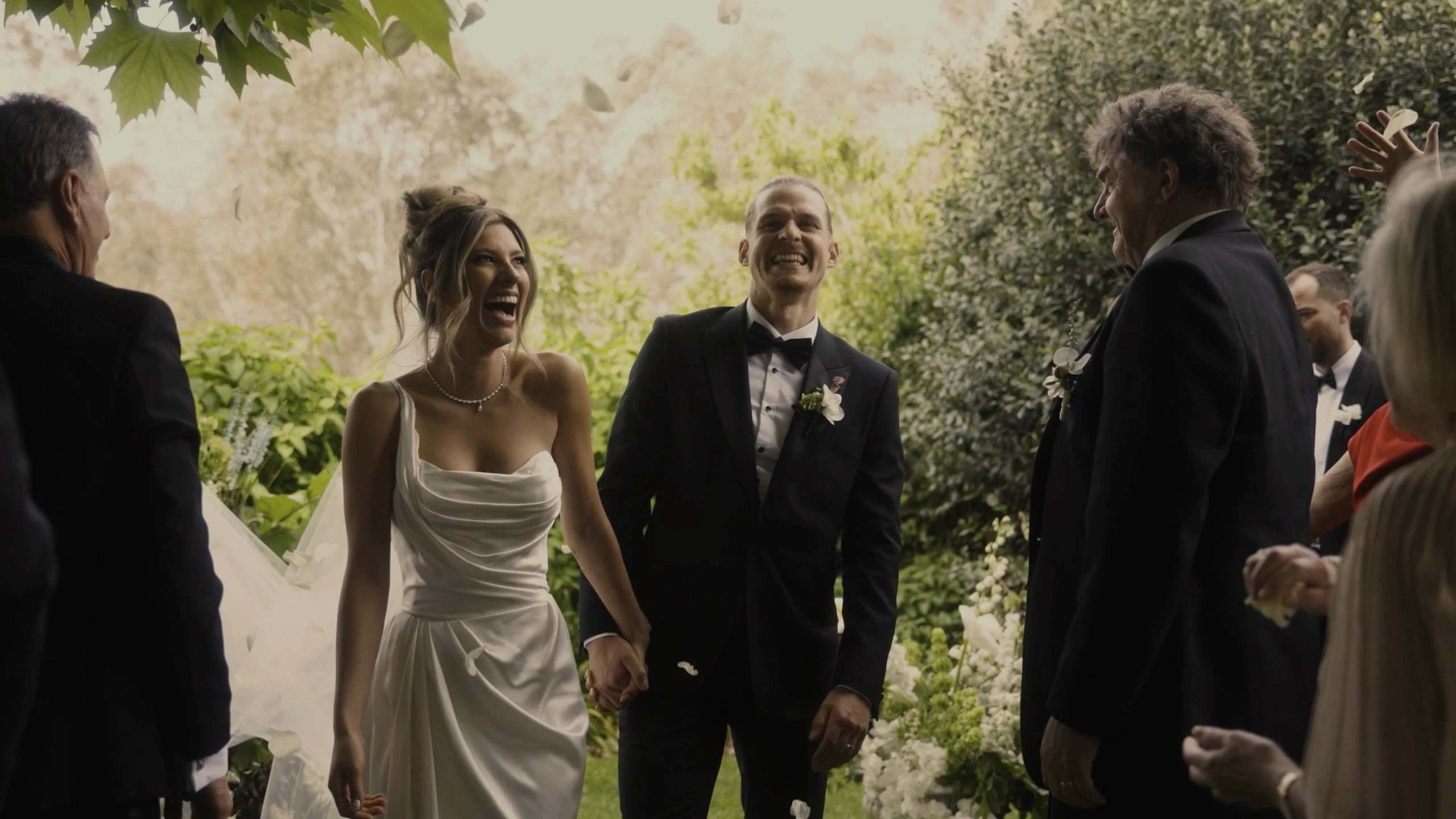 A joyful couple, elegantly dressed in a white wedding gown and black tuxedo, smiles radiantly as they walk hand in hand down a lush, outdoor garden aisle, surrounded by happy guests in formal attire.