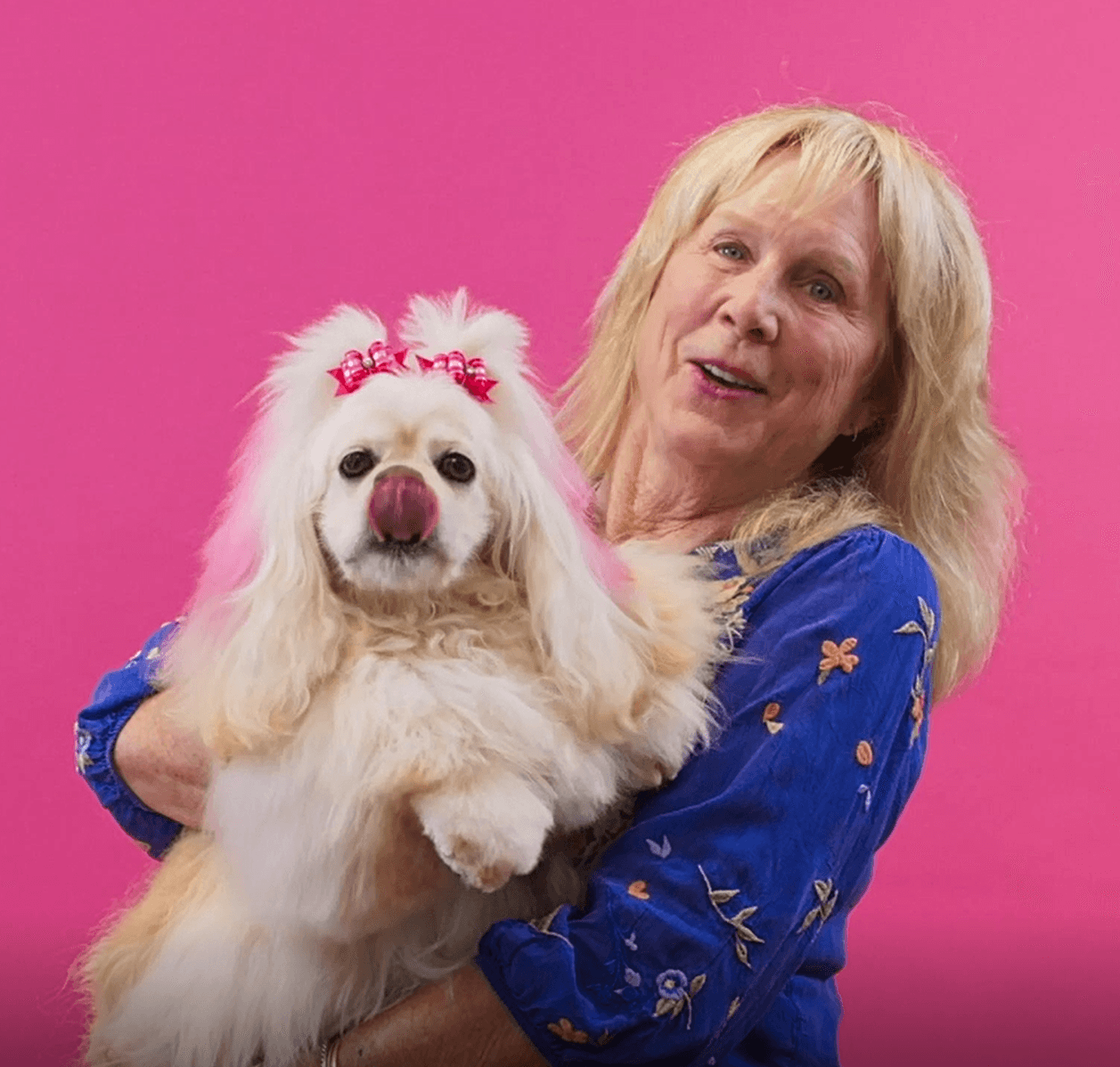 Smiling woman holding her fluffy white dog against a pink background to express love, care, and joyful grooming experience.