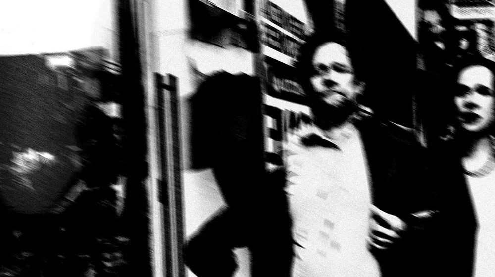 Blurry black-and-white street photograph of a man mid-motion, surrounded by harsh urban shadows and fragmented signage. High contrast and motion blur evoke a sense of anonymity, urgency, and voyeuristic tension.