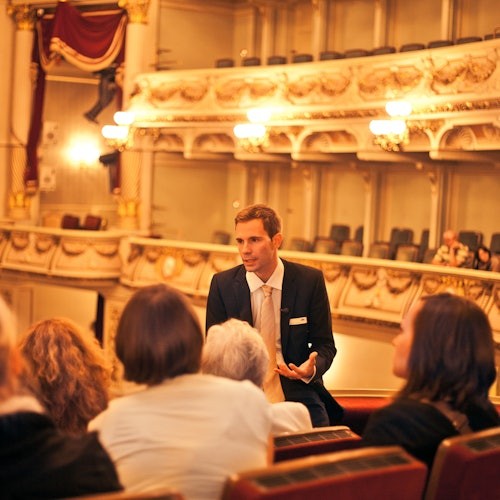 Mies puvussa puhuu pienelle joukolle ihmisiä koristeellisessa teatterissa, jossa on hienostunut valaistus ja sisustus.