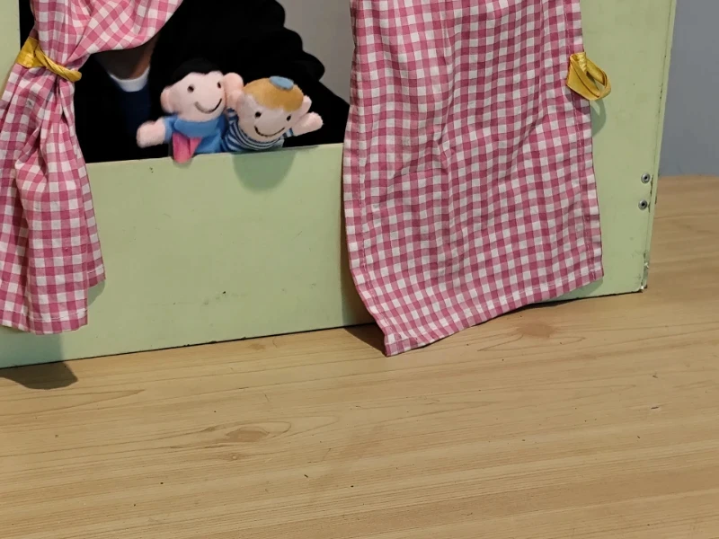 תיאטרון בובות לילדים עם וילונות משובצים בורדו-לבן ושתי בובות אצבע מחייכות על רקע שולחן עץ. בפעילות בקליניקה של סמדר עציץ נעים ולומדים
