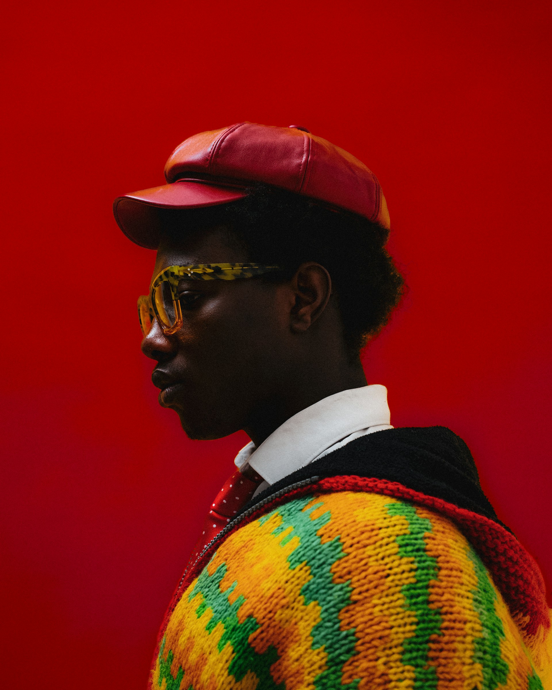 Side profile of a person wearing a red cap and yellow glasses against a red background.